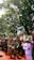 A group of people in traditional attire are participating in a procession under the shade of large trees. The participants appear to be involved in a cultural or ceremonial event, with some holding masks and decorative items.