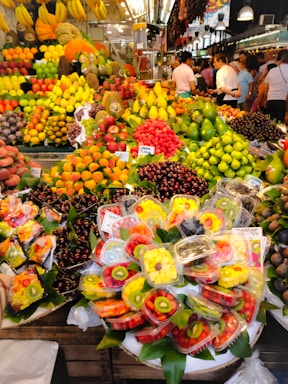 A vibrant and colorful fruit market stall is filled with an abundant variety of fresh fruits including bananas, grapes, strawberries, apples, mangoes, and cherries. Pre-packaged fruit trays containing sliced kiwis, strawberries, and pineapples are prominently displayed. The setting is bustling with people shopping, creating a lively atmosphere.