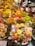 A vibrant and colorful fruit market stall is filled with an abundant variety of fresh fruits including bananas, grapes, strawberries, apples, mangoes, and cherries. Pre-packaged fruit trays containing sliced kiwis, strawberries, and pineapples are prominently displayed. The setting is bustling with people shopping, creating a lively atmosphere.