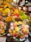 A vibrant and colorful fruit market stall is filled with an abundant variety of fresh fruits including bananas, grapes, strawberries, apples, mangoes, and cherries. Pre-packaged fruit trays containing sliced kiwis, strawberries, and pineapples are prominently displayed. The setting is bustling with people shopping, creating a lively atmosphere.