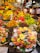 A vibrant and colorful fruit market stall is filled with an abundant variety of fresh fruits including bananas, grapes, strawberries, apples, mangoes, and cherries. Pre-packaged fruit trays containing sliced kiwis, strawberries, and pineapples are prominently displayed. The setting is bustling with people shopping, creating a lively atmosphere.