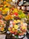 A vibrant and colorful fruit market stall is filled with an abundant variety of fresh fruits including bananas, grapes, strawberries, apples, mangoes, and cherries. Pre-packaged fruit trays containing sliced kiwis, strawberries, and pineapples are prominently displayed. The setting is bustling with people shopping, creating a lively atmosphere.