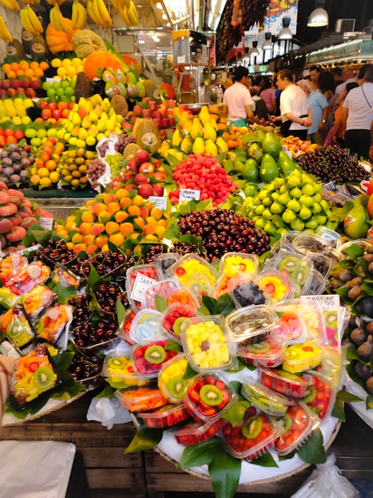 A vibrant and colorful fruit market stall is filled with an abundant variety of fresh fruits including bananas, grapes, strawberries, apples, mangoes, and cherries. Pre-packaged fruit trays containing sliced kiwis, strawberries, and pineapples are prominently displayed. The setting is bustling with people shopping, creating a lively atmosphere.