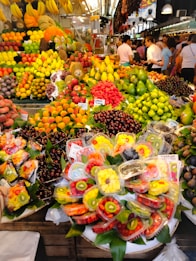 A vibrant and colorful fruit market stall is filled with an abundant variety of fresh fruits including bananas, grapes, strawberries, apples, mangoes, and cherries. Pre-packaged fruit trays containing sliced kiwis, strawberries, and pineapples are prominently displayed. The setting is bustling with people shopping, creating a lively atmosphere.