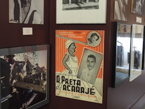 A collection of framed photographs and a colorful vintage poster are displayed on a wall. The poster features illustrations and black and white photos of two people, along with orange and cream stripes.