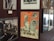 A collection of framed photographs and a colorful vintage poster are displayed on a wall. The poster features illustrations and black and white photos of two people, along with orange and cream stripes.