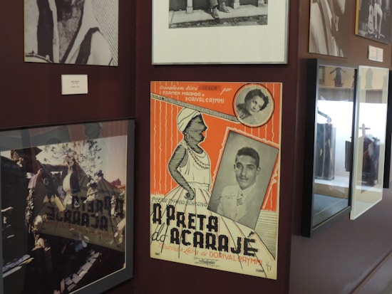 A collection of framed photographs and a colorful vintage poster are displayed on a wall. The poster features illustrations and black and white photos of two people, along with orange and cream stripes.
