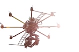 A drone with multiple rotors is flying against a bright white background. The structure of the drone is visible, showing intricate machinery and a mounted camera. Sunlight reflects off its metal parts, creating a lens flare effect.