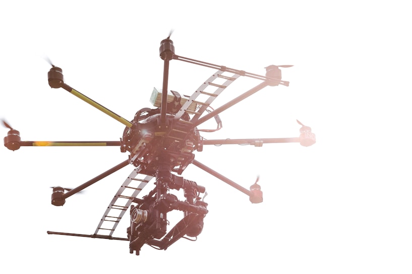A drone with multiple rotors is flying against a bright white background. The structure of the drone is visible, showing intricate machinery and a mounted camera. Sunlight reflects off its metal parts, creating a lens flare effect.