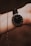 A wrist wearing a sleek, minimalist watch with a black dial and thin, white hour markers. The watch is secured with a black leather strap. A delicate gold chain bracelet is also visible, draped around the wrist, adding an elegant touch.