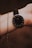 A wrist wearing a sleek, minimalist watch with a black dial and thin, white hour markers. The watch is secured with a black leather strap. A delicate gold chain bracelet is also visible, draped around the wrist, adding an elegant touch.