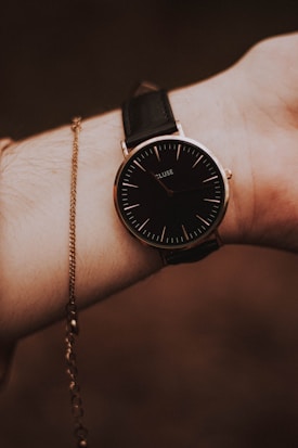 A wrist wearing a sleek, minimalist watch with a black dial and thin, white hour markers. The watch is secured with a black leather strap. A delicate gold chain bracelet is also visible, draped around the wrist, adding an elegant touch.