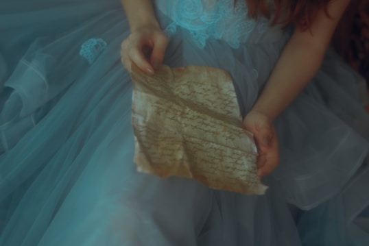 A person is holding an aged, parchment-like letter with handwritten text. They are dressed in a delicate, light blue gown with intricate lace details.