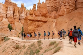 A group of hikers walks along a trail surrounded by striking rock formations featuring tall, reddish-orange hoodoos. The landscape is dry and rugged, with sparse vegetation visible. The hikers carry backpacks and are dressed in casual, outdoor attire suited for trekking.