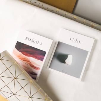 Two books are placed on a surface covered with decorative wrapping paper. The book on the left is titled 'Romans' and features a cover with abstract, earthy tones resembling sandstone layers. The book on the right is titled 'Luke' and has a minimalist cover with a single white object against a muted background. The wrapping paper beneath them has a white base with a gold geometric pattern.