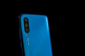A close-up of a blue smartphone with multiple rear cameras aligned vertically on the side. The phone has a modern design with smooth edges, and the words '48 MP AI CAMERA' are visible near the camera module. The background is completely black, highlighting the phone's sleek and glossy finish.