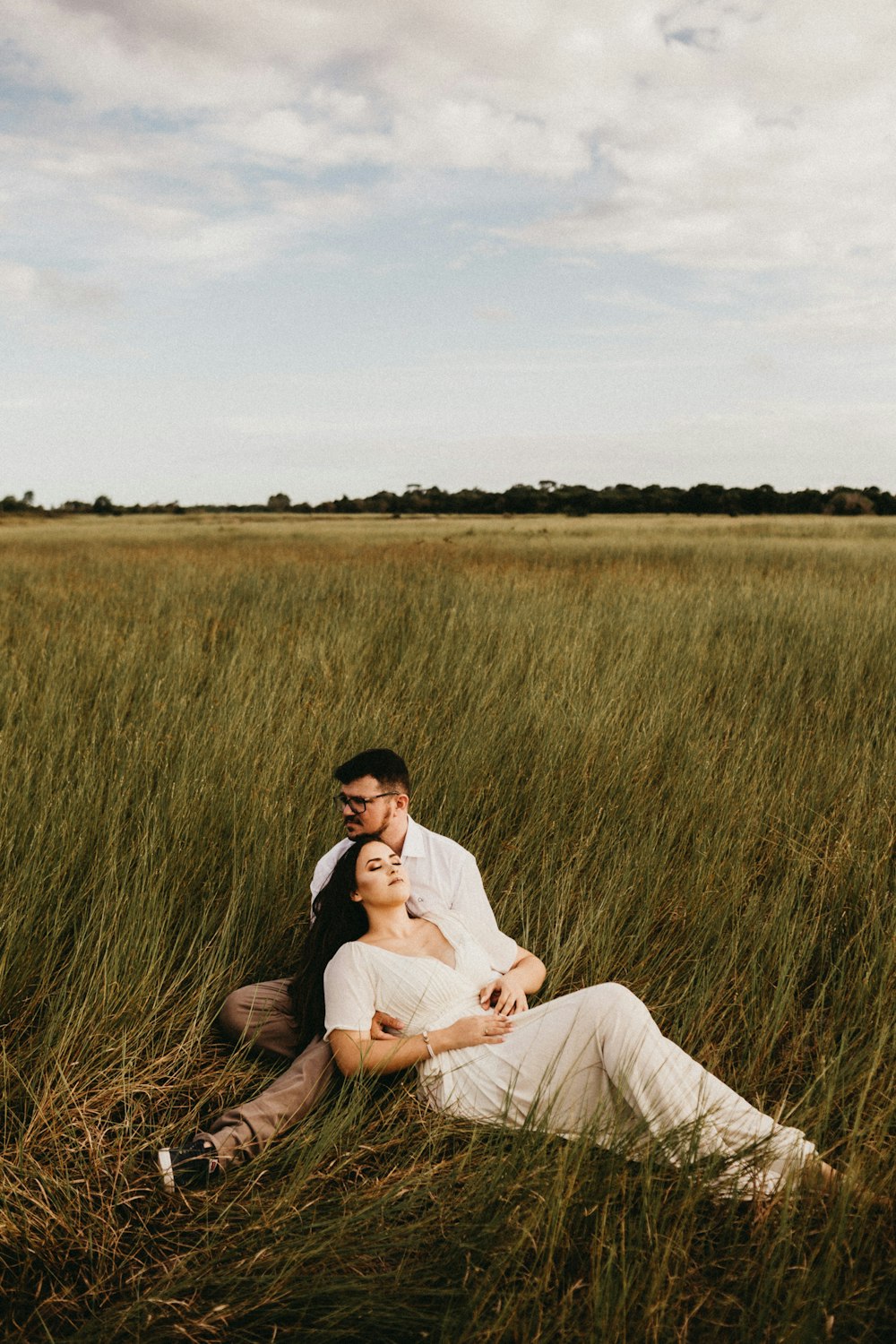 man wearing white dress shirt sitting on grass field and woman wearing white dress laying on man