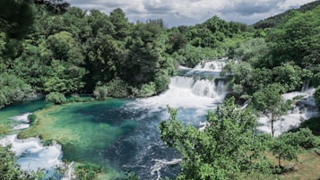 A lush, green landscape with a cascading waterfall surrounded by dense trees and foliage. The water flows over multiple levels, creating white frothy waves as it descends into a tranquil pool below. The scene is vibrant and rich, with a mix of clear blue and emerald green hues in the water, contrasting with the deep greens of the forest.