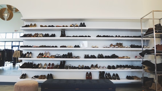 Shelves filled with various types of shoes are neatly arranged in a retail environment. The footwear includes sneakers, high heels, and formal shoes displayed on multiple rows. The room features a modern, minimalist design with other garments visible on racks to the left and additional items on a metal shelf to the right.