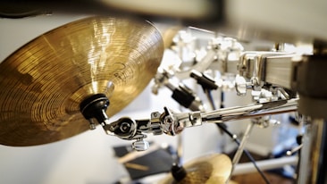 A close-up of a drum kit focusing on a cymbal with a shiny, metallic finish. The hardware is polished and chrome-plated, reflecting light, and creating a sleek, modern look. The background is slightly blurred, adding depth and focus to the musical instruments.