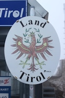 An oval-shaped sign with an intricate illustration of a heraldic eagle holding crossed branches, crowned with a decorative wreath. The words 'Land Tirol' are prominently displayed above and below the eagle. The sign is mounted on a pole, and a blue sign with the word 'Tirol' is partially visible in the background. The overall setting appears misty or foggy.