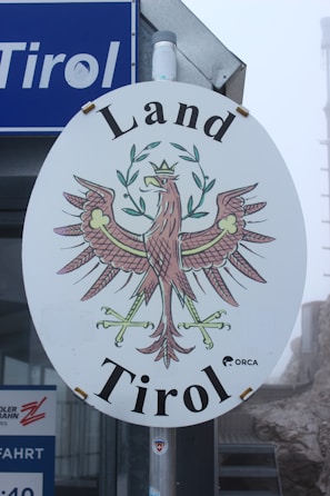 An oval-shaped sign with an intricate illustration of a heraldic eagle holding crossed branches, crowned with a decorative wreath. The words 'Land Tirol' are prominently displayed above and below the eagle. The sign is mounted on a pole, and a blue sign with the word 'Tirol' is partially visible in the background. The overall setting appears misty or foggy.