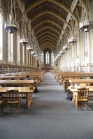A grand library hall with a high, vaulted ceiling featuring large hanging lamps. The room is lined with tall windows and bookshelves along the walls. Wooden study tables and chairs are neatly arranged in rows, with a few people seated and studying.