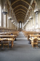 A grand library hall with a high, vaulted ceiling featuring large hanging lamps. The room is lined with tall windows and bookshelves along the walls. Wooden study tables and chairs are neatly arranged in rows, with a few people seated and studying.