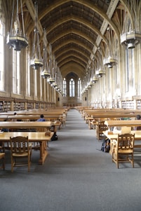 A grand library hall with a high, vaulted ceiling featuring large hanging lamps. The room is lined with tall windows and bookshelves along the walls. Wooden study tables and chairs are neatly arranged in rows, with a few people seated and studying.