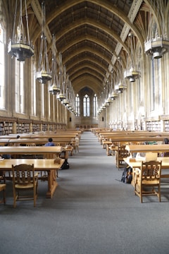 A grand library hall with a high, vaulted ceiling featuring large hanging lamps. The room is lined with tall windows and bookshelves along the walls. Wooden study tables and chairs are neatly arranged in rows, with a few people seated and studying.