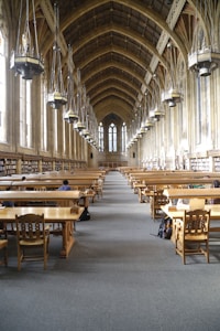 A grand library hall with a high, vaulted ceiling featuring large hanging lamps. The room is lined with tall windows and bookshelves along the walls. Wooden study tables and chairs are neatly arranged in rows, with a few people seated and studying.