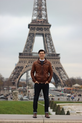 A person stands confidently in front of the Eiffel Tower, wearing a brown leather jacket and jeans. The scene is set in a bustling area with people and vehicles in the background. The sky is overcast, creating a muted atmosphere.