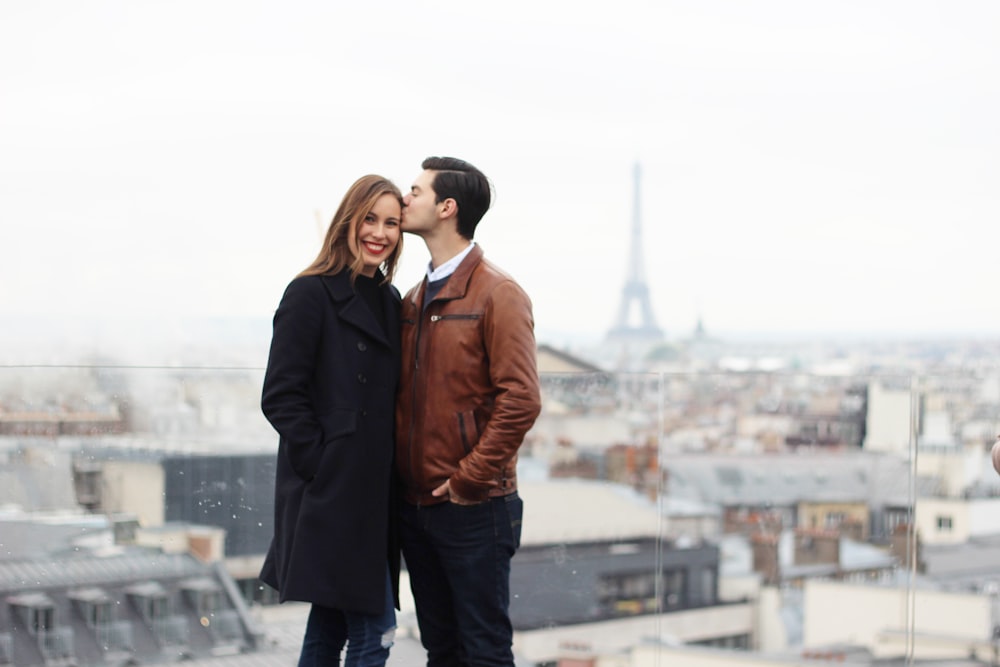 man wearing brown leather jacket and black jeans kissing forehead of woman wearing black trench coat