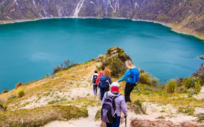 A group of hikers is descending a trail overlooking a beautiful turquoise lake surrounded by steep, rocky hills. The landscape is lush with patches of greenery, and wildflowers dot the path. The hikers are dressed in outdoor gear and are carrying backpacks, adding to the sense of an adventurous journey.