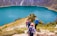 A group of hikers is descending a trail overlooking a beautiful turquoise lake surrounded by steep, rocky hills. The landscape is lush with patches of greenery, and wildflowers dot the path. The hikers are dressed in outdoor gear and are carrying backpacks, adding to the sense of an adventurous journey.