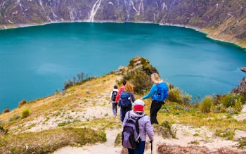 A group of hikers is descending a trail overlooking a beautiful turquoise lake surrounded by steep, rocky hills. The landscape is lush with patches of greenery, and wildflowers dot the path. The hikers are dressed in outdoor gear and are carrying backpacks, adding to the sense of an adventurous journey.