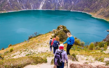 A group of hikers is descending a trail overlooking a beautiful turquoise lake surrounded by steep, rocky hills. The landscape is lush with patches of greenery, and wildflowers dot the path. The hikers are dressed in outdoor gear and are carrying backpacks, adding to the sense of an adventurous journey.