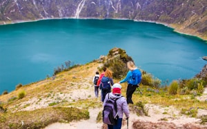 A group of hikers is descending a trail overlooking a beautiful turquoise lake surrounded by steep, rocky hills. The landscape is lush with patches of greenery, and wildflowers dot the path. The hikers are dressed in outdoor gear and are carrying backpacks, adding to the sense of an adventurous journey.