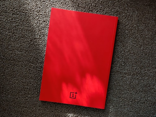 A red notebook rests on a textured gray carpet, with lighting that casts soft shadows and highlights on its surface. The notebook features a small logo at the bottom center.