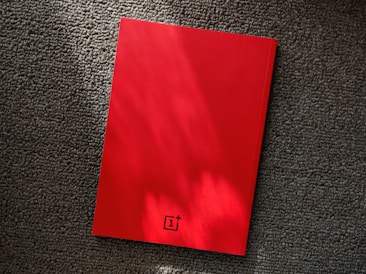 A red notebook rests on a textured gray carpet, with lighting that casts soft shadows and highlights on its surface. The notebook features a small logo at the bottom center.