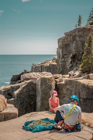 A coastal cliffside with several people engaged in rock climbing activities. Two individuals are prominently seen preparing gear and ropes, wearing climbing helmets. The background features the ocean and rugged cliffs, with some trees on the cliff tops.