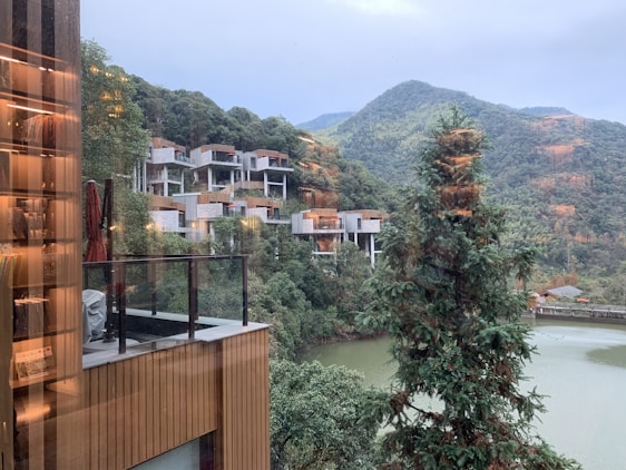 A serene landscape featuring modern hillside structures surrounded by lush greenery. The buildings are constructed on a mountain slope, with a reflective glass surface in the foreground possibly showing interior lights from within a building. In the distance, forested hills and a river or lake can be seen.