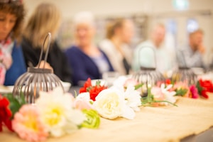 A table decorated with colorful flowers and lanterns, set in a soft-focus background where several people are sitting. The flowers include varieties of pink, red, white, and some greens, while the metallic lanterns add a rustic element. The scene suggests a gathering or a casual event.
