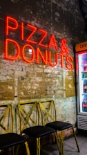 A neon sign displaying the words 'Pizza & Donuts' illuminates a rustic wall with partially exposed bricks and faded paint. Below the sign, there are three yellow metal chairs with black seats. To the right, a refrigerator stocked with drinks is partially visible.