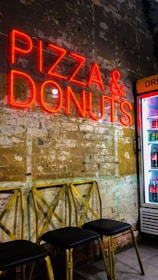 A neon sign displaying the words 'Pizza & Donuts' illuminates a rustic wall with partially exposed bricks and faded paint. Below the sign, there are three yellow metal chairs with black seats. To the right, a refrigerator stocked with drinks is partially visible.