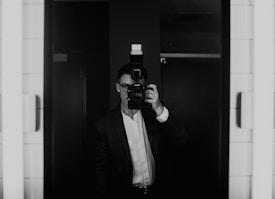 A person in formal attire stands in front of a mirror, holding a camera with a flash attachment, obscuring their face. The scene is captured in black and white, providing a classic and introspective atmosphere.