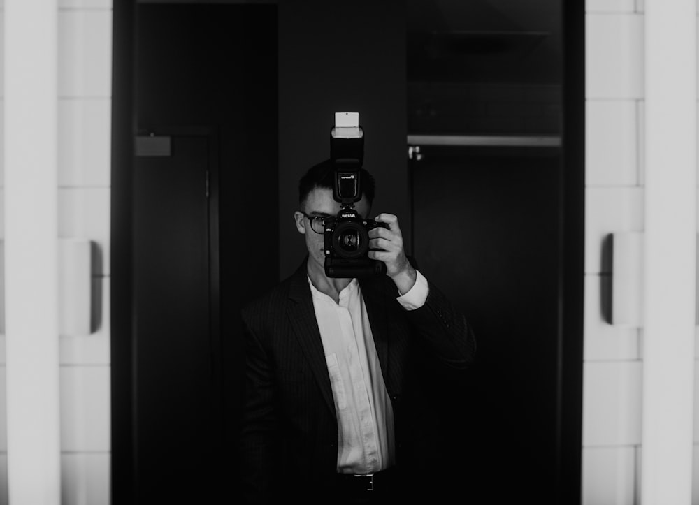 grayscale photo of man wearing suit holding DSLR camera