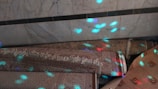 A group of rolled-up carpets with intricate patterns lies on the floor against a marble wall. The surface of the carpets is illuminated by colorful light patterns, predominantly in blue and pink hues.