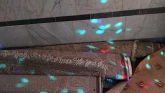 A group of rolled-up carpets with intricate patterns lies on the floor against a marble wall. The surface of the carpets is illuminated by colorful light patterns, predominantly in blue and pink hues.