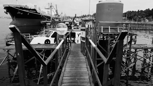 A dock extends into a body of water with several boats moored alongside, including a large ship named 'Grace Poseidon'. The wooden pier leads towards the water, bordered by railings. A large cylindrical container is situated on the pier, while industrial structures and more vessels are visible in the background.